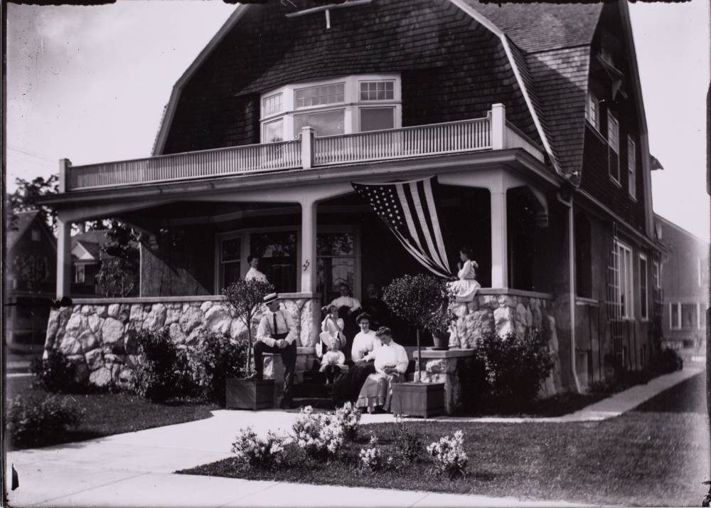 Black and white antique photo of family sitting on covered front porch of two story home